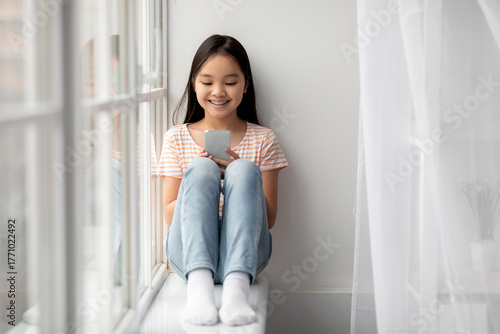 A young girl sits comfortably by a large window, smiling as she uses her smartphone. The sunlight brightens the cozy space, creating a cheerful atmosphere for her leisure time. Free Download A young girl sits comfortably by a large window, smiling as she uses her smartphone. The sunlight brightens the cozy space, creating a cheerful atmosphere for her leisure time. - GFXLoad
