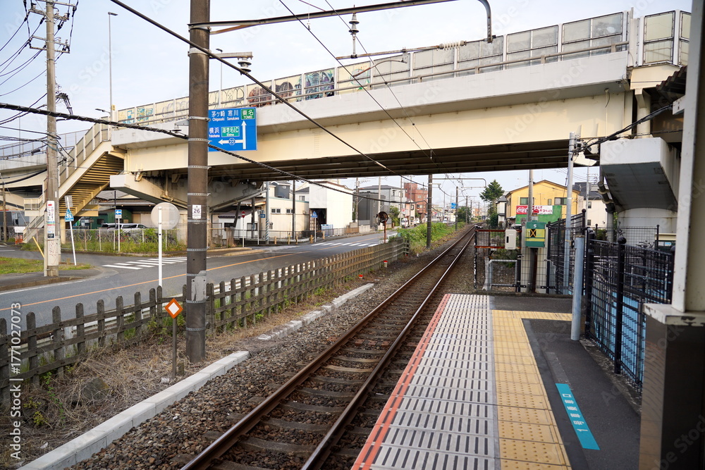 Fototapeta premium 住宅地の踏切の脇にあるローカル線の線路とホームの風景です。頭上には歩道橋や架線があり日本の都市近郊の日常の交通インフラが感じられる情景です。