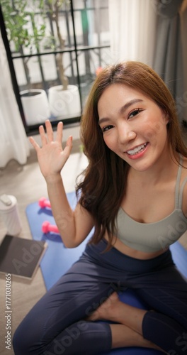 POV young Asian woman in sportswear live streaming fitness session on social media, smiling and waving at the camera. Female health influencer interacting with followers or teaching online workout.