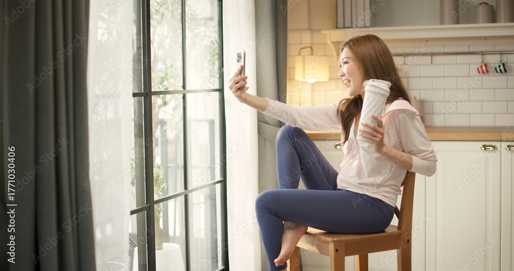 Obraz premium Young Asian woman in sportswear holds a protein shake bottle taking selfie with smartphone for social media after home workout. Healthy lifestyle, fitness motivation, self care, post-workout recovery.