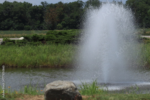 Small Fountain with Water Splashes and Droplets against Green Grass and Trees
