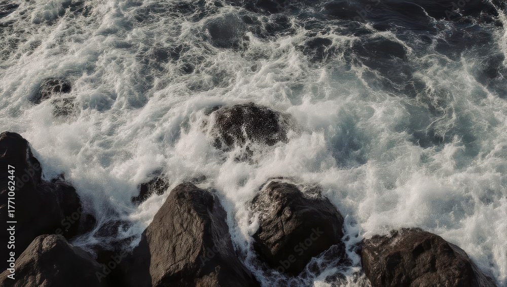 Obraz premium Crashing ocean waves churn against dark, jagged rocks, creating white foam and motion
