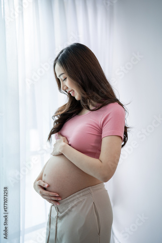Free Download Asian young pregnant woman gently holds and strokes her belly in house. - GFXLoad