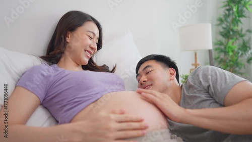 Asian husband and pregnant wife lying together on bed in cozy bedroom. 