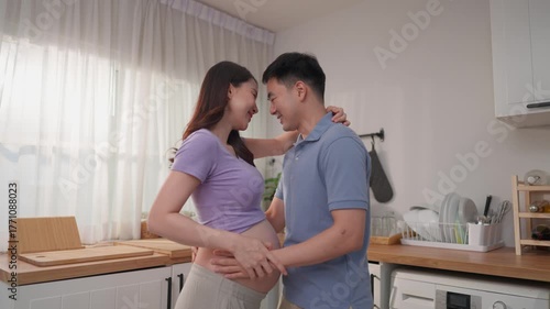 Asian husband and pregnant wife standing together in kitchen at home.