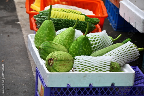 Green melons in white foam box with mesh protection, orange basket with yellow-netted melons behind, blue plastic basket base, outdoor gray pavement, market produce display, Taiwan.