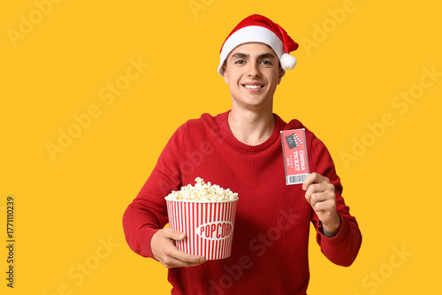 Young man in Santa hat with cinema ticket and bucket of popcorn on yellow bac...