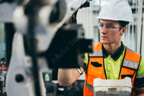 Male engineer in safety gear inspecting and controlling remote control at industrial robotic arm, Workplace safety, maintenance, and skilled industrial labor in a factory technology