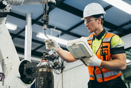 Male technician in hardhat and vest operating a robotic welding arm with a handheld control panel. Industrial automation. Skilled industrial maintenance at the robot factory