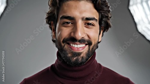 Smiling man with brown hair and a beard wearing a maroon turtleneck sweater against a gray backdrop