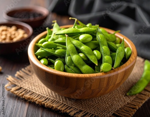 Rustic wooden bowl of fresh green edamame on woven mat with soybeans and dark sauce in background.