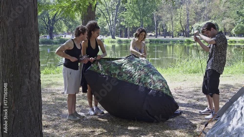 Four happy friends prepare their outdoor shelter by a serene lake in a green park. They work together, enjoying a summer day setting up for a fun picnic.