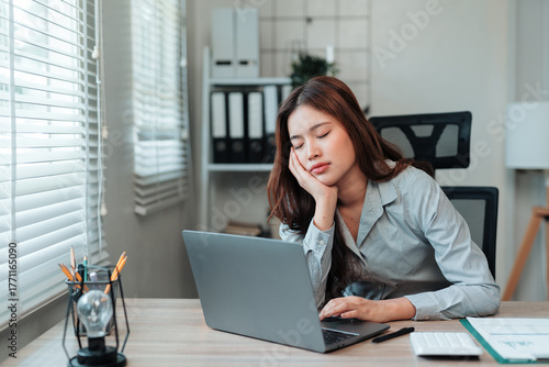 Exhausted young Asian woman sleeping at office desk with laptop. Experiencing burnout, fatigue, and lack of sleep