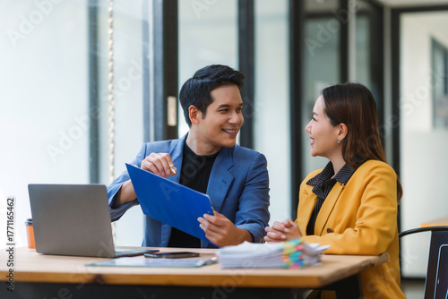 Two Asian businesspeople reviewing documents and discussing work, showing successful teamwork and professional collaboration in a modern office setup