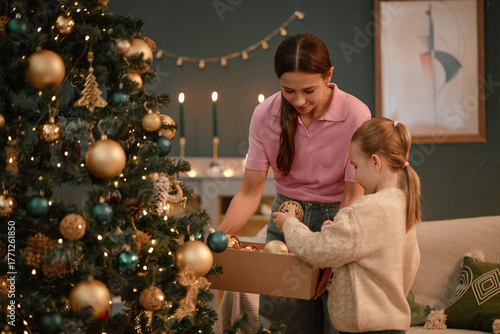 Caucasian young woman helping child girl decorating Christmas tree with ornaments, both smiling and interacting near festive decorations in cozy living room