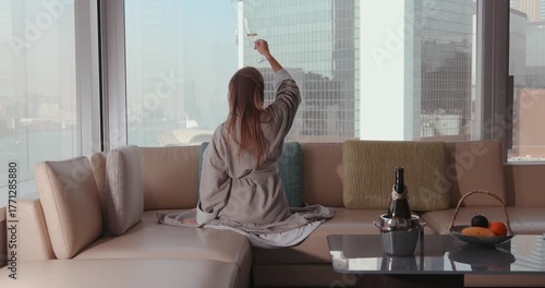 Woman in luxury hotel suite enjoying morning relaxation in bathrobe with champagne, city view, and breakfast fruit basket.