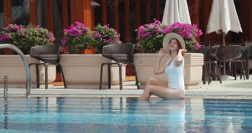 Woman relaxing by outdoor pool, wearing sunhat and swimsuit, surrounded by lush flowers and patio furniture. Summer leisure, vacation, resort lifestyle concept.
