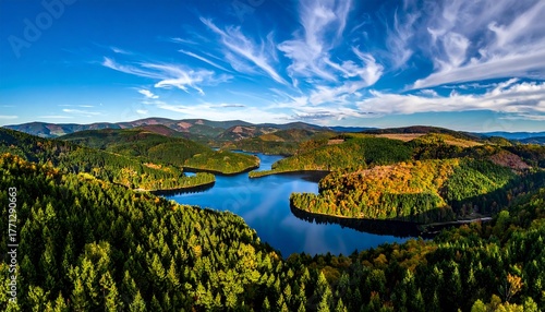 Lake surrounded by lush autumn forests, vibrant hues, and wispy clouds beneath a vivid blue sky creates a scenic vista