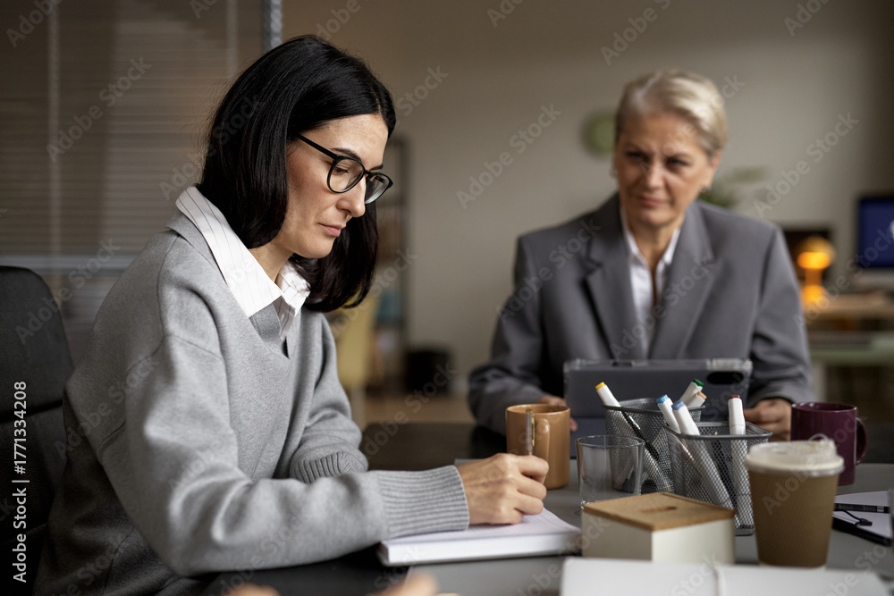 Fototapeta premium Young adult Caucasian woman writing in notebook while sitting at desk with middle aged Caucasian woman in background holding digital tablet during business meeting