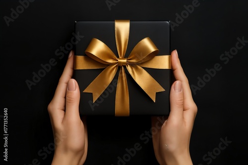 Luxury Christmas photo of hands holding black gift box with golden satin bow on dark background, elegant festive composition