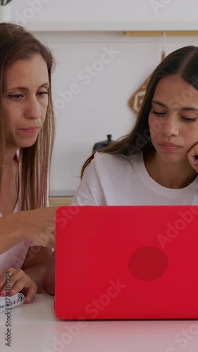 Vertical video. Mid adult mother is supporting her 15-year-old teenage daughter with school homework at the kitchen table while using a laptop. Education, family support, and home learning concept.