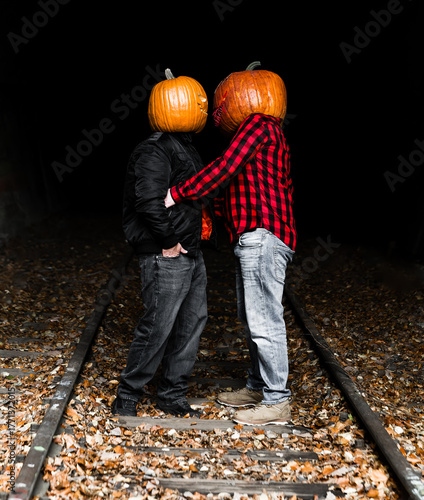 Couple wearing pumpkin heads share a playful moment on a leaf-covered train track in autumn twilight