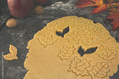 Shortcrust pastry being cut into autumn leaf shapes on a floured dark surface, surrounded by colorful fall leaves — cozy seasonal cookie preparation and festive baking process.