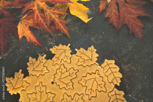 Shortcrust pastry being cut into autumn leaf shapes on a floured dark surface, surrounded by colorful fall leaves — cozy seasonal cookie preparation and festive baking process.