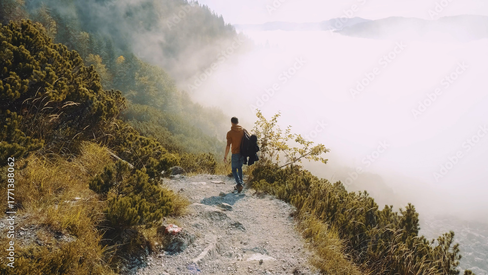 Obraz premium Man walks along a misty mountain trail surrounded by lush greenery and stunning views during early morning light