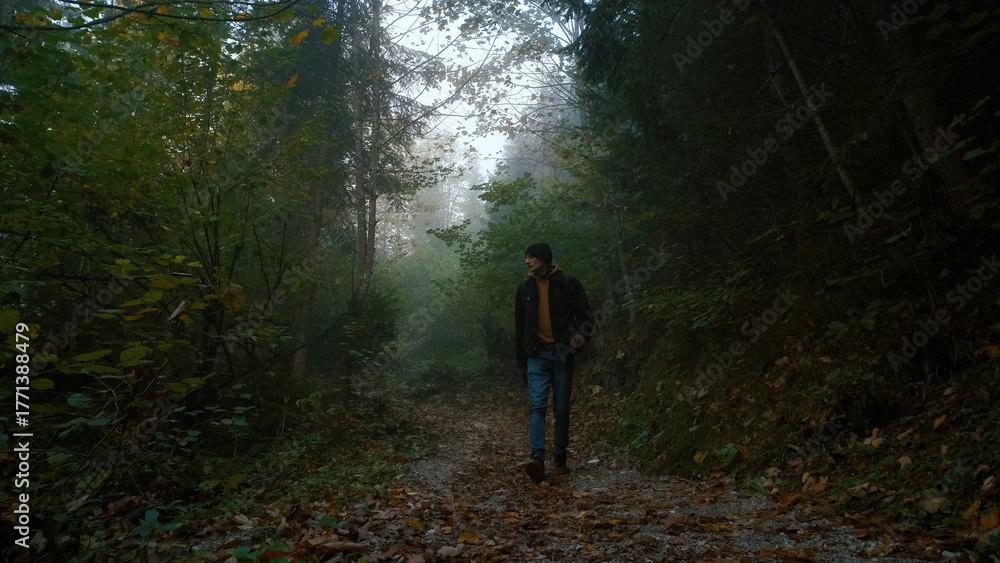 Obraz premium Man walking through a foggy forest pathway surrounded by trees and autumn leaves on a quiet day