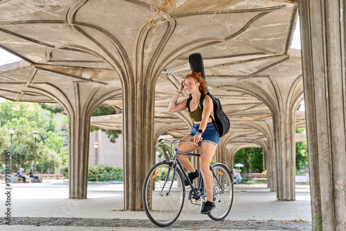 Bild auf Leinwand Young musician riding bicycle carrying guitar in city park