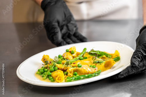 Wallpaper Mural A person wearing black gloves presents a white oval plate of pasta with green beans and peas in a creamy yellow sauce, garnished with microgreens Torontodigital.ca
