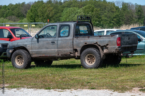 Old pickup truck parked in an overgrown field near a car lot on a sunny day
