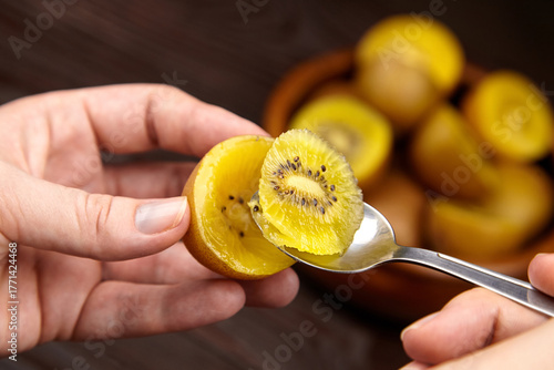 Close-up of hands holding a spoon with a slice of yellow kiwi fruit, showcasing its vibrant color and texture against a wooden background with whole fruits