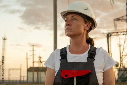 Female electrical worker wearing hard hat at a power plant looks toward horizon at sunset