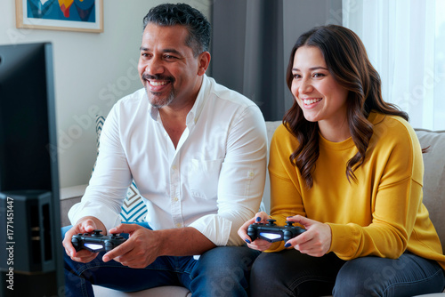 Woman and Hispanic middle aged man sitting on sofa playing video game together using wireless controllers smiling and looking at television screen in modern living room