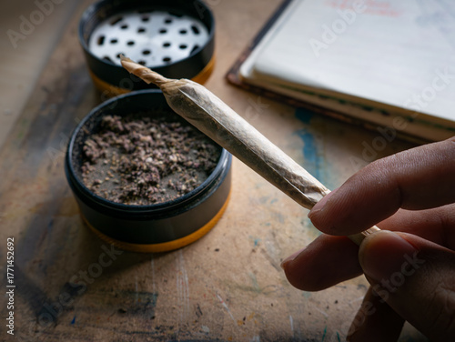 Hand holding rolled marijuana joint with grinder and cannabis herb on wooden table for recreational use