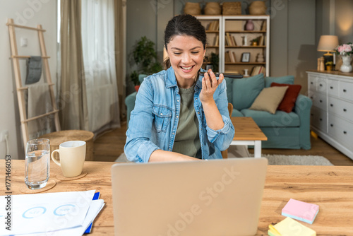 Freelance businesswoman in home office reviewing charts and dictating a voice note while analyzing financial reports on a laptop — remote work, strategy planning productivity and tech-driven workflow