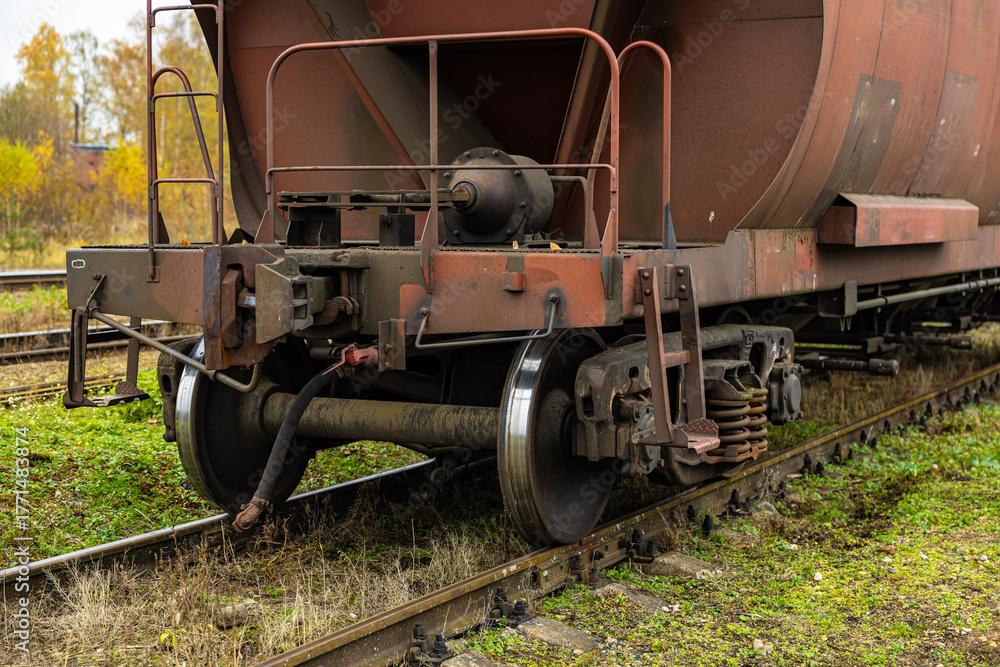 Naklejka premium Side view of an old, rusted freight wagon showing its undercarriage, coupling mechanism, suspension, and metal ladder, positioned on overgrown railway tracks