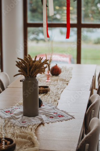 Autumnal coziness: a vase of ears of corn on a wooden table decorated with a linen tablecloth and embroidery. A warm, homely atmosphere, rustic style, and natural harmony.