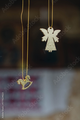 Two Christmas tree decorations - a golden angel and a rocking horse - hang on thin strings against a blurred background.
