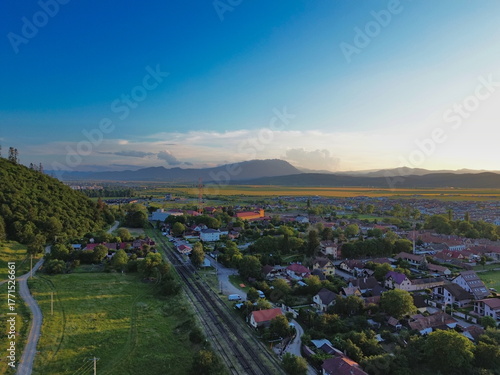 Aerial view of train tracks slicing through the vibrant green landscape and quaint rooftops, nestled against a backdrop of distant mountains at sunset, Cristian, BraÈ™ov, Romania.