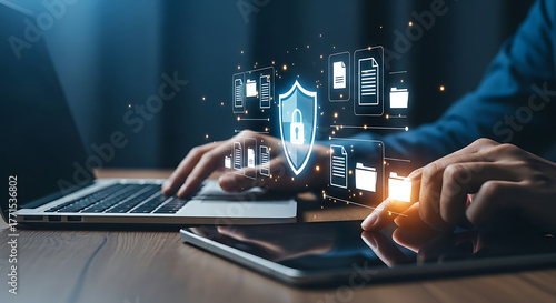 Person working on a laptop and a tablet with a shield padlock icon and file icons, representing data security, protection, and cybersecurity