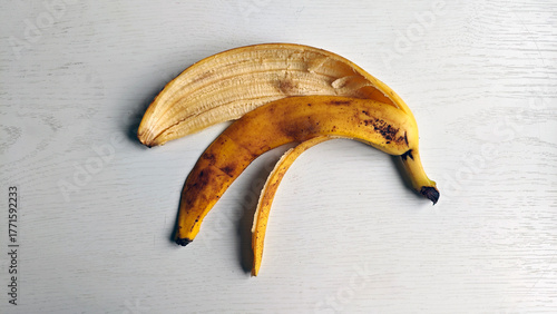 Peeled banana skin lying on white wooden surface. Banana peel has some brown spots, indicating ripe. Banana peel is curved and has yellow color with some brown spots. Organic garbage.