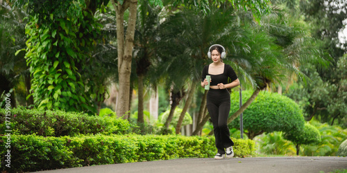 Young woman jogging in a lush park with headphones, enjoying a refreshing drink while staying active and healthy in natural surroundings