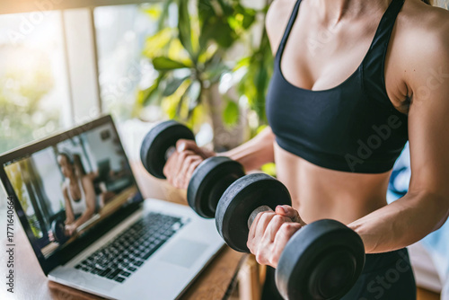 Close-up of hands gripping dumbbells, laptop with trainer blurred in background, soft sunlight, muted green plant, highlighting muscle engagement and exercise detail.