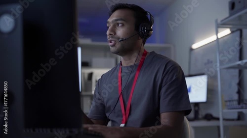 Remote work online technical support concept. Portrait young adult indian man wearing headset typing keyboard working in office. It support business customer service technology.