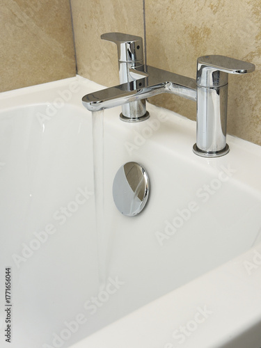 Closeup of water flowing from a pair of hot and cold, shiny chrome lever taps, which are positioned over the end of a white bath tub.