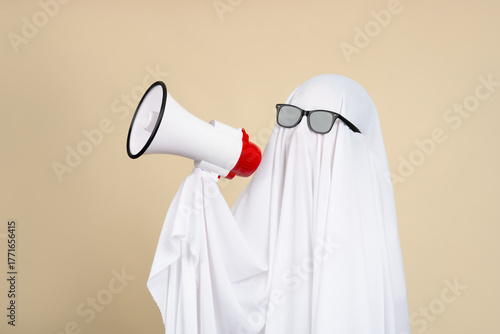 Funny Halloween ghost wearing white cloth shouting into megaphone isolated on beige background. Happy Halloween or weird concept.