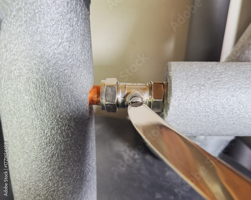 Closeup of an isolating valve between lagged indoor pipes, with the rounded end of a cutlery knife being used to turn a screw, to open or shut off the water supply.